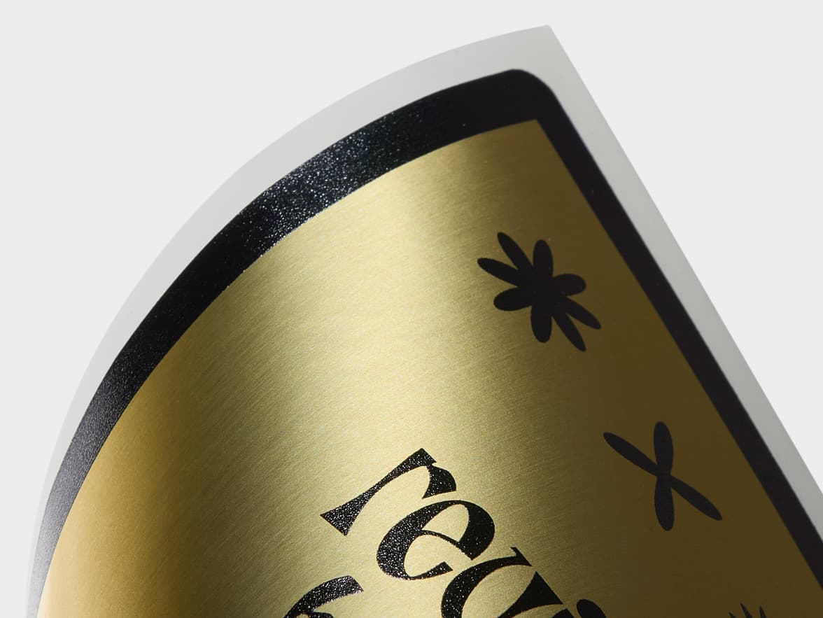 A close up image of the corner of a gold metallic rectangle sticker with black customization.